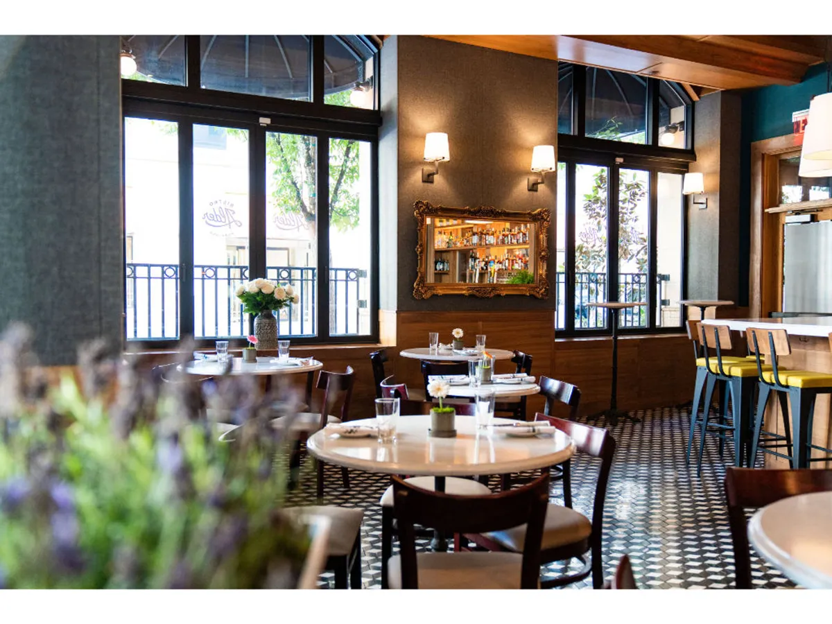A bright and welcoming space featuring tables set for dining, large windows flooding the room with natural light, a decorative mirror, and a bar area in the background. This setting exudes warmth and hospitality, inviting guests to enjoy meals or socialize at any time.