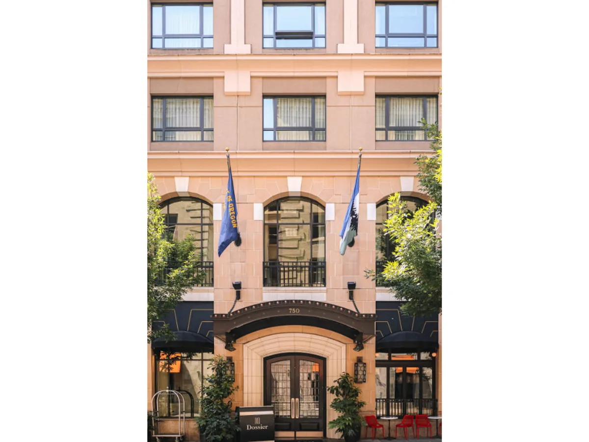 An exterior view of the hotel's entrance archway, adorned with flags and unique window designs, surrounded by lush landscaping. The scene captures people arriving, socializing in the outdoor seating area, and vibrant city life around the building, all bathed in beautiful morning light.