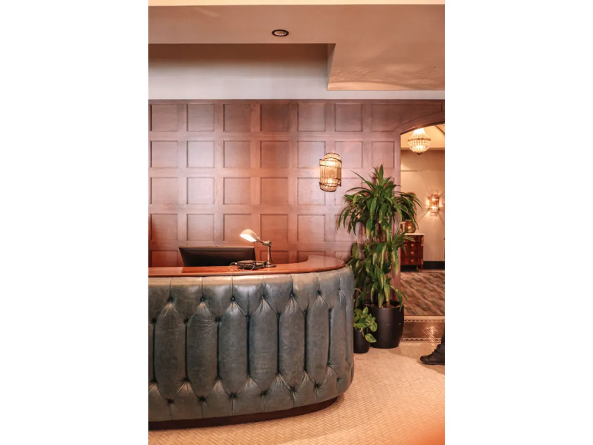 A stylish, curved reception desk sits front and center, framed by elegant wood paneling and modern hanging light fixtures. An indoor plant softens the space, and the overall mood is warm and inviting—signaling a polished, hotel-level welcome and attentive guest service.