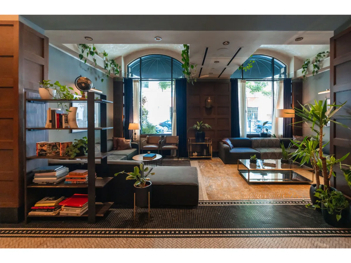 The scene showcases a living area bathed in natural light, with large windows, comfortable seating, lush greenery, and a decorative bookshelf. There's a clear sense of a multi-functional space that encourages both relaxation and productivity.