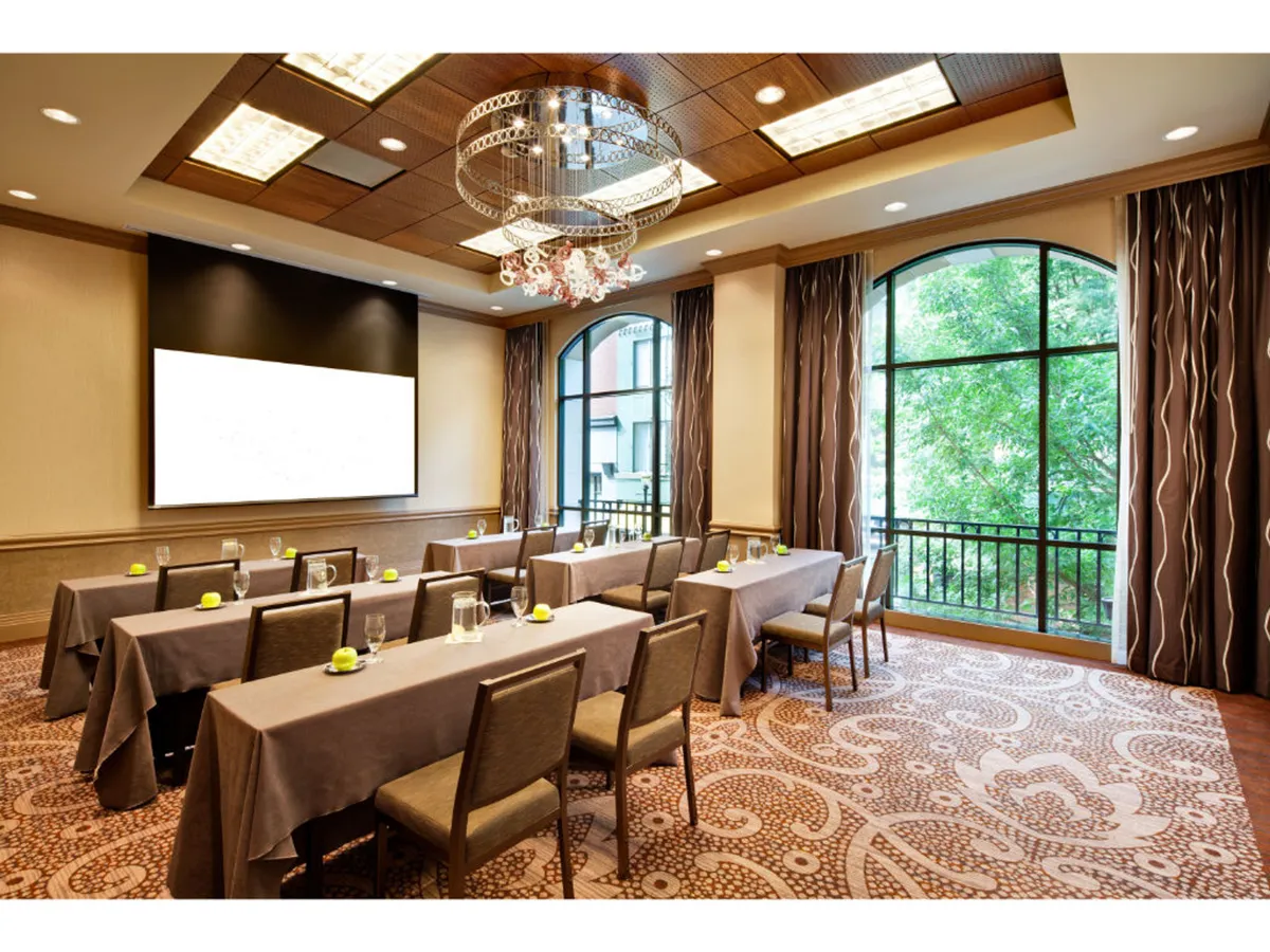 A modern meeting room featuring tables arranged for collaborative sessions, a large screen for presentations, an elegant chandelier providing soft lighting, and expansive windows allowing natural light to flood the space. The view through the windows offers a subtle connection to the city outside, creating a blend of work focus and urban energy.