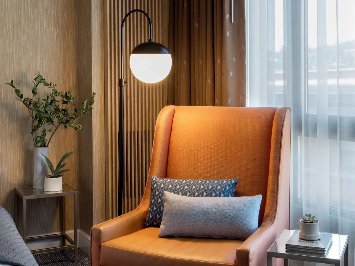 This image features a cozy orange armchair paired with a floor lamp, decorative pillows, a plant, and a side table, evoking a tranquil and comfortable in-room atmosphere. The space looks ideal for relaxing, reading, or enjoying a fresh cup of coffee.