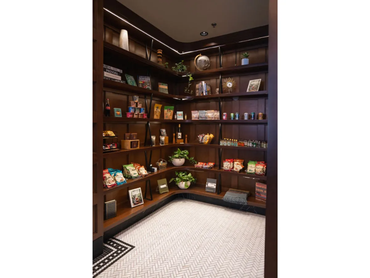 A set of wooden shelves are filled with a variety of products, including snacks and beverages, accented by decorative plants and unique decor items. The scene is welcoming and suggests easy access to refreshments and local treats.