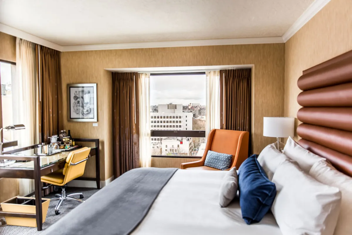A corner of the bedroom featuring a king-sized bed with decorative pillows, a cozy orange accent chair, and a work desk. The space is bathed in natural light with a spectacular city view visible through the windows, emphasizing privacy and comfort.