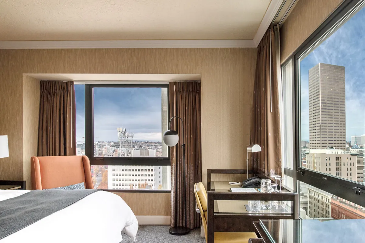 The image showcases a sunlit hotel room corner in Portland, featuring expansive windows that frame sweeping city views. A plush, inviting bed is perfectly placed to take in the skyline, while a modern desk offers a stylish spot for work or relaxation. The space exudes a calming atmosphere, ideal for unwinding after exploring the city or enjoying a productive moment with inspiring vistas.