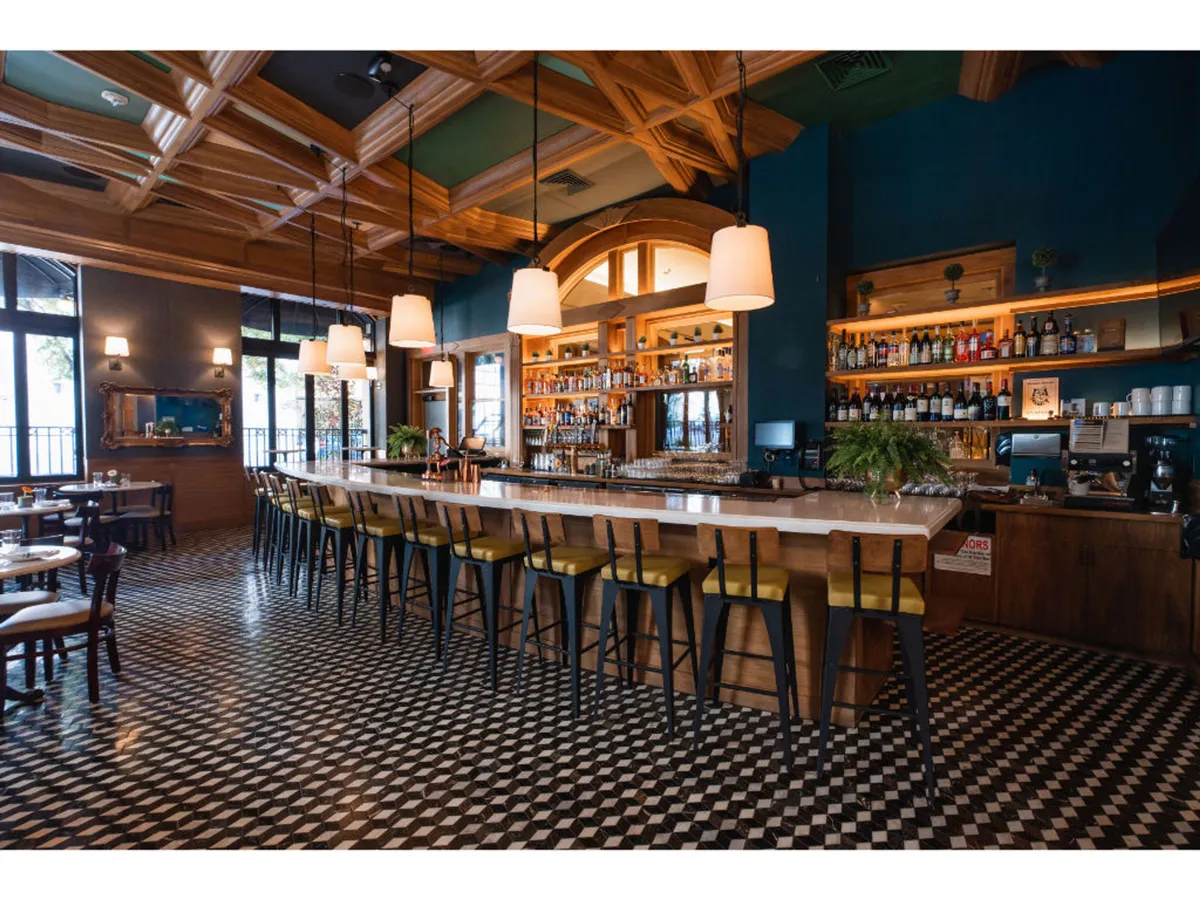 A lively scene inside a stylish bar, featuring a long bar with high stools, a decorative wooden ceiling, and shelves lined with bottled spirits. The atmosphere is bustling and energetic, suggesting socializing and enjoying craft cocktails.