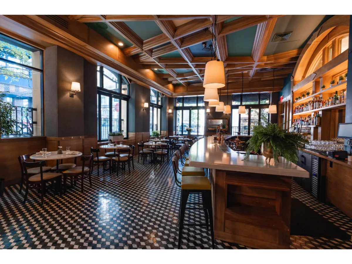 A welcoming bar and dining area with a stylish bar counter, comfortable seating, warm chandeliers, and a lively yet relaxed atmosphere. The bar is well-stocked, hinting at craft cocktails and social opportunities after a busy workday.