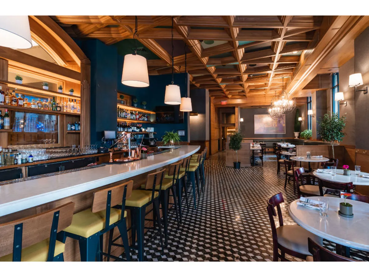 A sophisticated and cozy interior scene featuring the Bar Alder area, with elegant chandeliers, a decorative ceiling, tables adorned with plants, and guests enjoying craft cocktails and gourmet dishes in a lively environment.